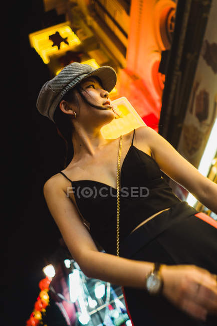 Elegante jovem mulher asiática bonita de pé na rua iluminada à noite e olhando para longe — Fotografia de Stock