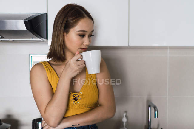 Вдумчивая молодая женщина с чашкой, стоящей на кухне — стоковое фото