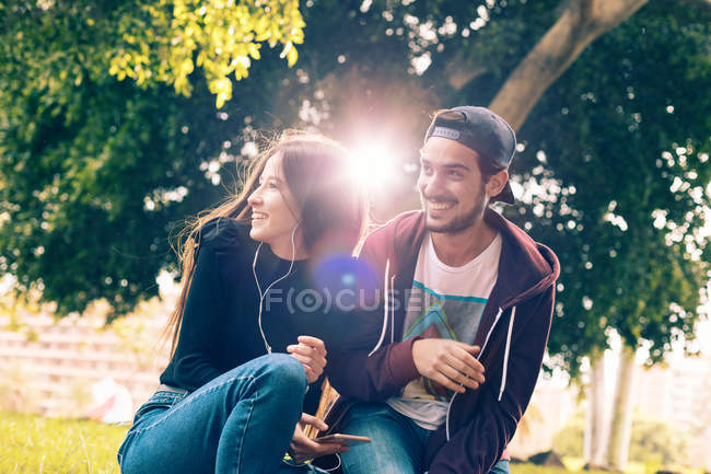 Сміючись молода пара сидить з смартфон в парку — стокове фото