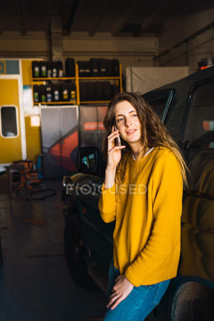 Woman talking on smartphone while leaning on car in garage — Stock Photo