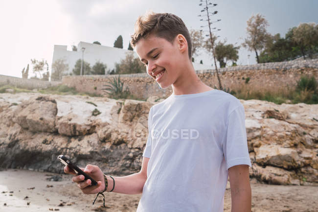 Menino com o smartphone sorrindo enquanto está de pé na costa rochosa — Fotografia de Stock