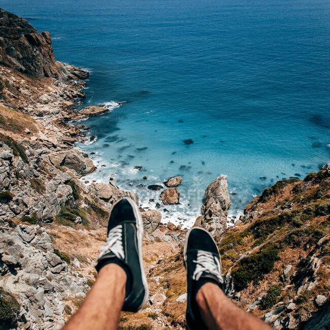 Cultivo piernas de turista irreconocible sentado en el acantilado en el océano azul. - foto de stock