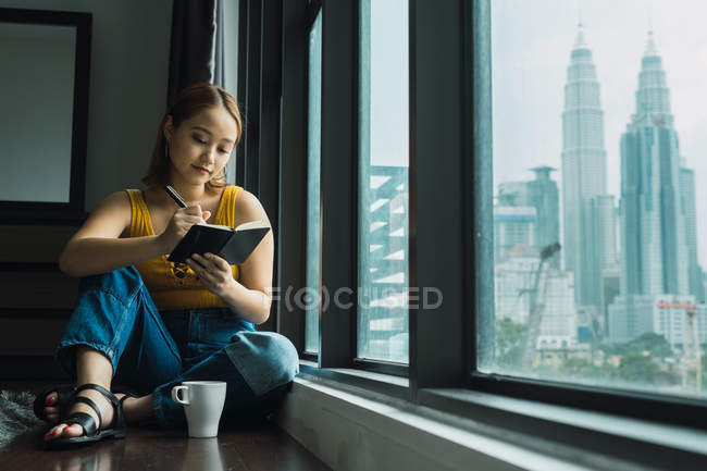 Женщина с чашкой сидит на полу у окна и делает заметки — стоковое фото
