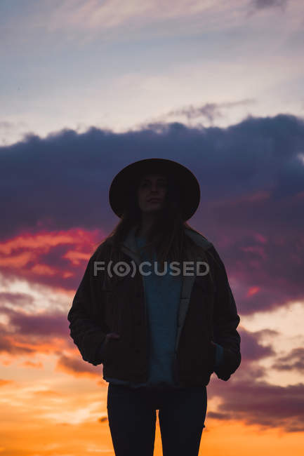 Thoughtful woman in hat and jacket standing at sunset under dramatic sky — Stock Photo