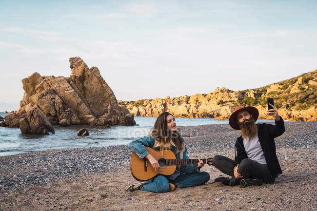 Молодая женщина, сидящая на пляже и играющая на гитаре для мужчины с мобильного телефона — стоковое фото