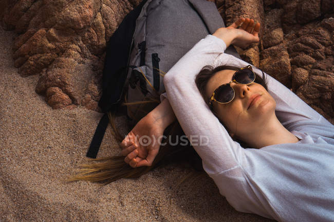 Туристична жінка в сонцезахисних окулярах лежить на рюкзаку на піску — стокове фото