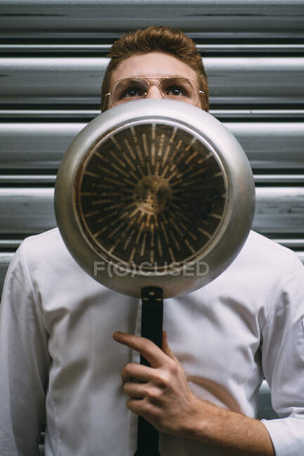 Pensive young man in glasses and white jacket covering face with frying pan looking up. — Stock Photo