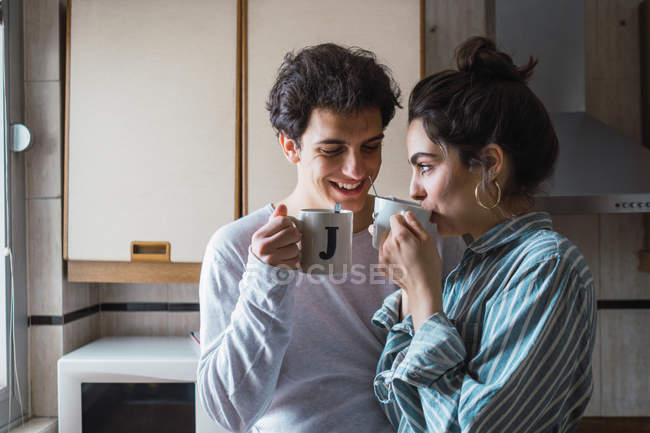 Щаслива молода пара стоїть з двома чашками і посміхається на кухні вранці — стокове фото