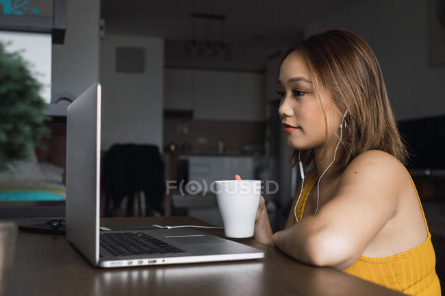 Mulher com copo usando laptop à mesa no apartamento — Fotografia de Stock