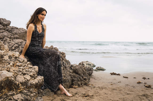 Retrato de mulher sorridente em vestido sentado na rocha no mar — Fotografia de Stock