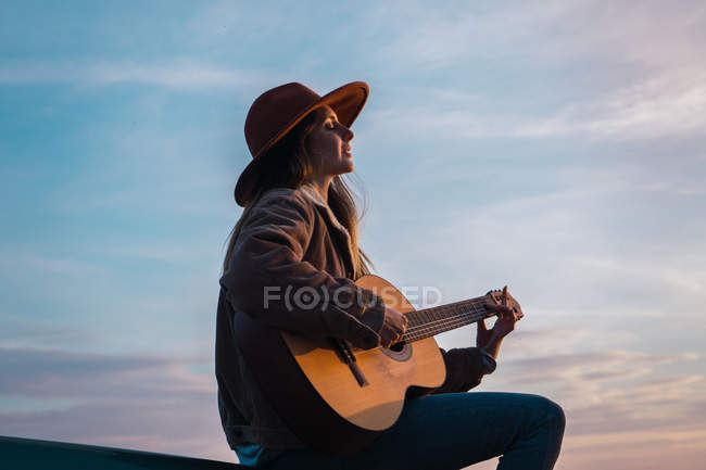 Donna seduta sulla macchina e suonare la chitarra al tramonto — Foto stock