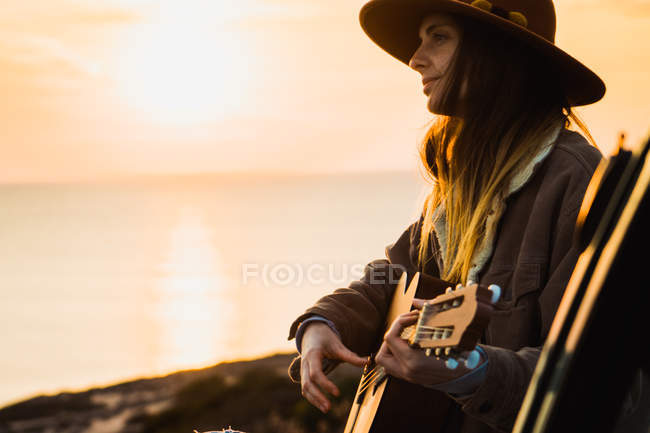 Mulher tocando guitarra à beira-mar ao pôr do sol — Fotografia de Stock