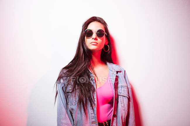 Досить молода рожева одягнена жінка в сонцезахисних окулярах стоїть на білій стіні і дивиться на камеру — стокове фото