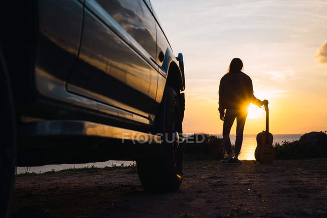 Donna in piedi in auto e tenendo la chitarra al mare al tramonto — Foto stock