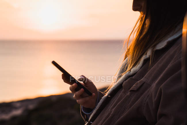 Frau benutzt Smartphone, während sie bei Sonnenuntergang an der Küste steht — Stockfoto