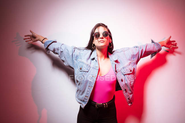 Pretty young pink dressed woman in sunglasses standing at white wall with arms outstretched — Stock Photo