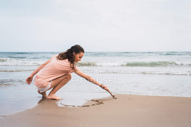 Девочка-подросток, приседающая и рисующая палкой на песке на берегу моря — стоковое фото