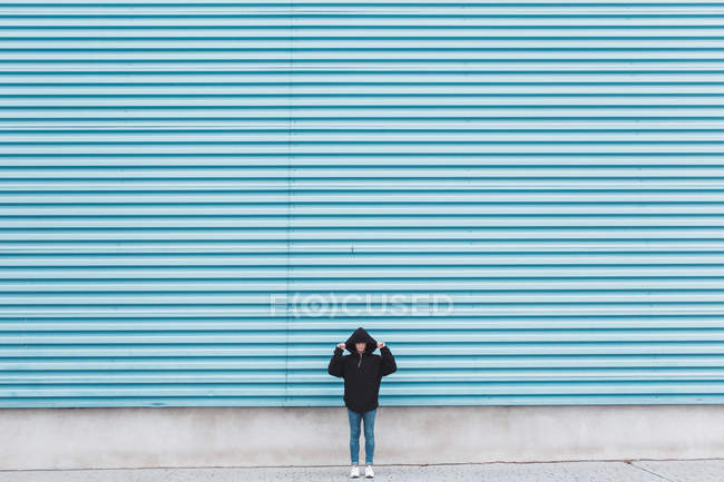 Homem de casaco com capuz em pé contra a parede de metal azul — Fotografia de Stock