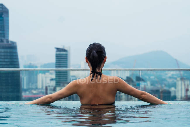 Mujer asiática relajándose en la piscina con vista a la ciudad en el fondo - foto de stock