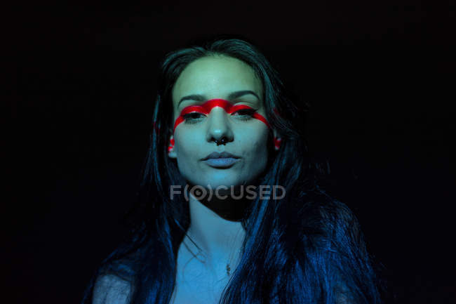 Jeune femme attrayante avec ligne rouge sur le visage en regardant la caméra sur fond noir — Photo de stock