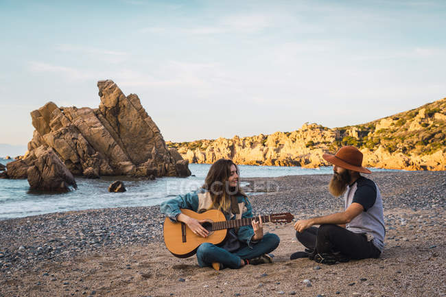 Pretty woman sitting on beach and playing guitar for man — Stock Photo