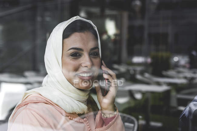 Mujer marroquí con hijab y vestido árabe tradicional hablando por teléfono detrás del cristal de la ventana - foto de stock
