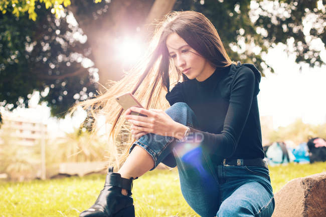 Jeune femme assise sur le rocher et utilisant un smartphone dans le parc — Photo de stock