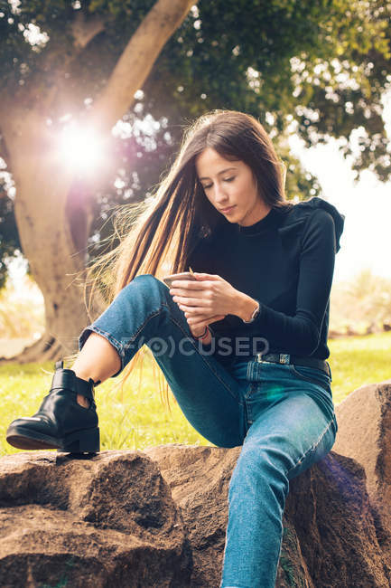 Giovane donna seduta sulla roccia e utilizzando smartphone nel parco — Foto stock