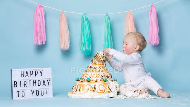 Lindo niño sentado en el pastel de cumpleaños y comiendo sobre fondo azul - foto de stock