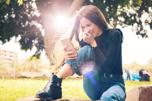 Jovem mulher rindo sentado no rock e usando smartphone no parque — Fotografia de Stock