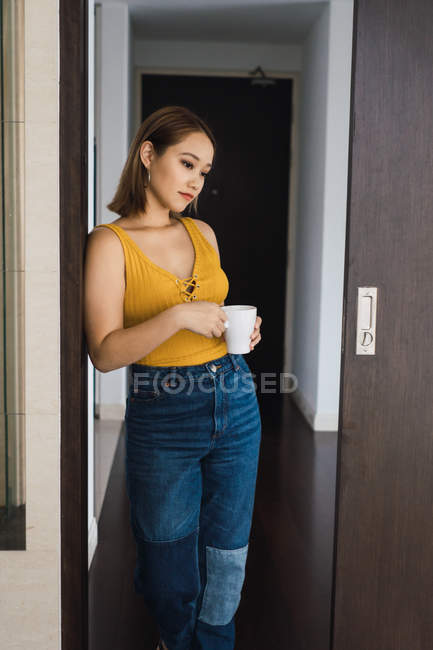 Стомлена молода жінка з чашкою спирається на стіну вдома — стокове фото