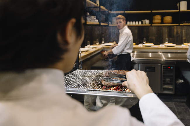 Chef preparando carne asada en el restaurante con colega en el fondo - foto de stock