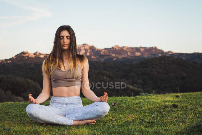 Молода жінка в спортивному одязі сидить на зеленому газоні і робить йогу з закритими очима — стокове фото