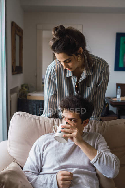 Вдумлива молода жінка дивиться на хлопця, який п'є з чашки на дивані — стокове фото