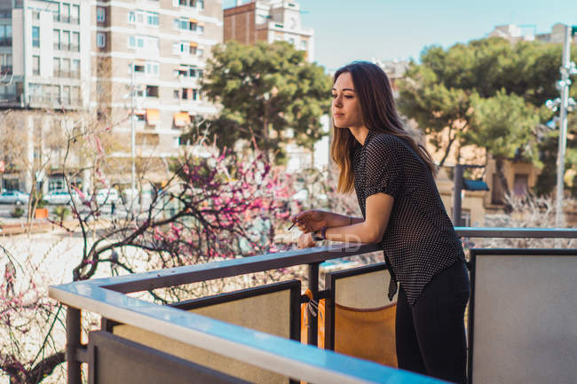 Verträumte junge Frau steht und raucht auf Balkon in der Stadt — Stockfoto