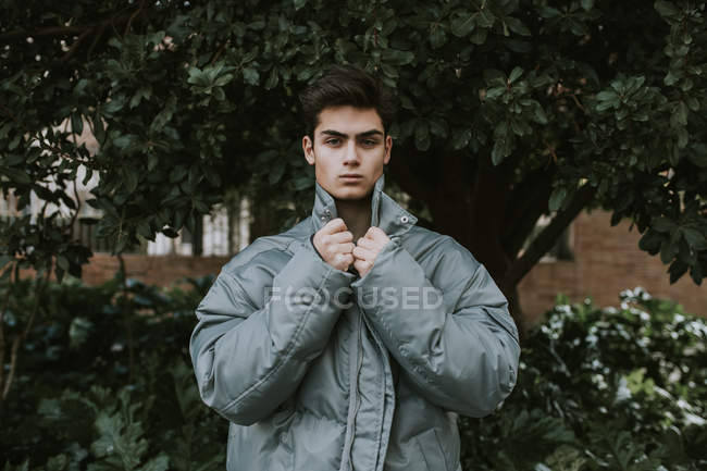 Giovane bel adolescente in grigio elegante giacca calda in piedi davanti all'albero verde — Foto stock