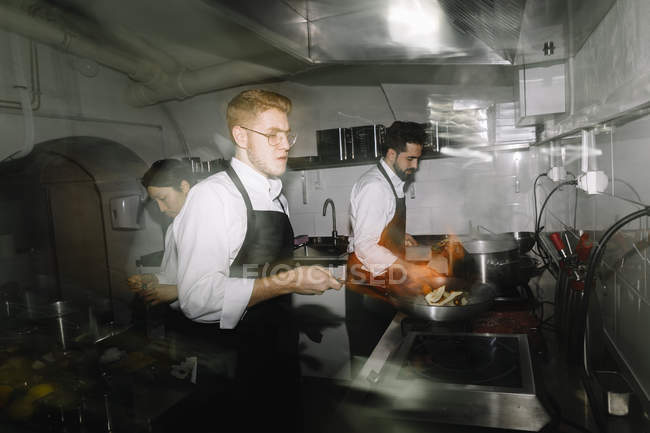Cook making a flambe in restaurant kitchen with colleagues on background — Stock Photo