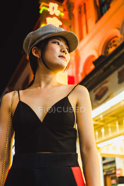 Giovane donna asiatica in piedi su strada illuminata di notte e guardando altrove — Foto stock