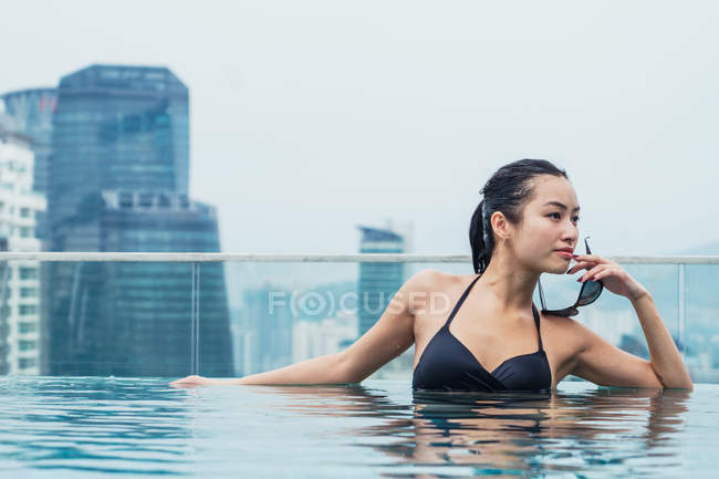 Mujer asiática relajándose en la piscina con rascacielos modernos en el fondo - foto de stock