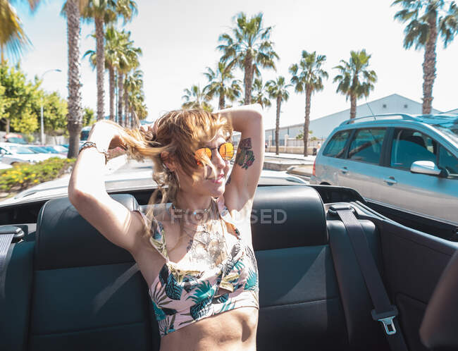 Усміхнена жінка в сонцезахисних окулярах і короткий літній одяг, що їде на пасажирському сидінні в кабріолеті з руками в повітрі на дорозі з пальмами навколо — стокове фото
