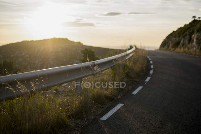 Захисний паркан і асфальтована дорога на заході сонця назад освітлені в горах . — стокове фото