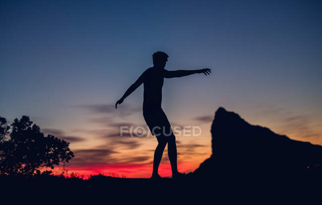 Silueta de persona irreconocible de pie junto a la roca en las luces del atardecer. - foto de stock