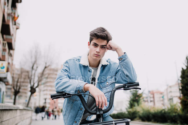 Jovem adolescente bonito inclinado no guidão da bicicleta e olhando para a câmera na rua — Fotografia de Stock