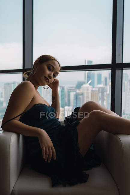 Bella donna in abito nero rilassante in poltrona in appartamento con vista sulla città — Foto stock