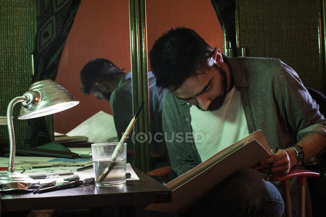 Adulto artista homem sentado à mesa e desenho em grande caderno de esboços em casa. — Fotografia de Stock