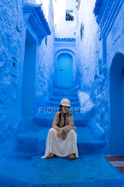 Mujer sonriente sentada en las escaleras de la ciudad marroquí teñida de azul - foto de stock