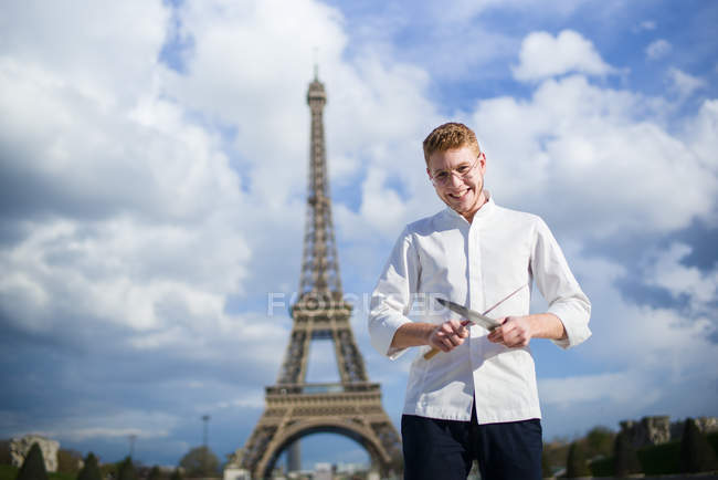 Улыбающийся рыжеволосый повар с ножами, стоящий перед Эйфелевой башней в Париже — стоковое фото