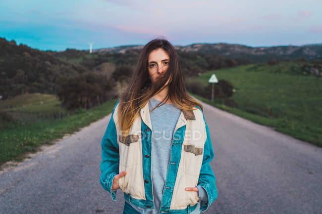 Jovem mulher em roupas casuais em pé na estrada na natureza e olhando para a câmera — Fotografia de Stock