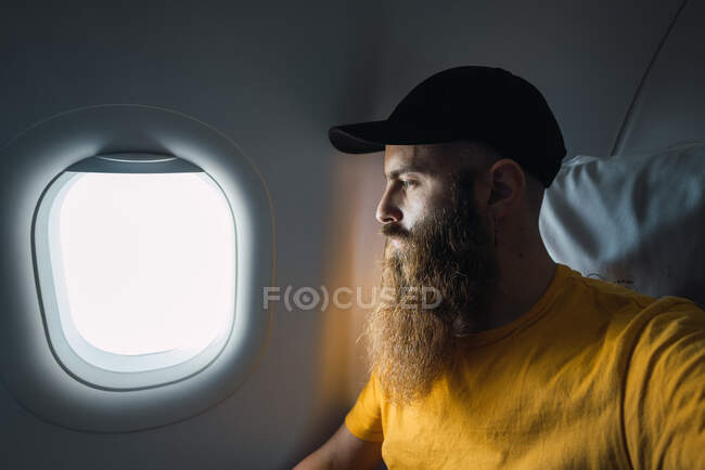Hombre adulto casual con barba usando gorra y camiseta amarilla mirando hacia otro lado en la ventana mientras toma vuelo en avión - foto de stock