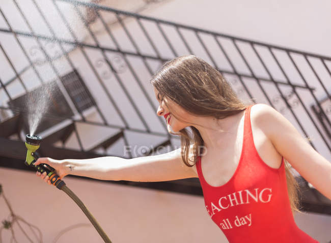 Young pretty woman in red swimsuit having fun in backyard with spraying water hose in sunlight — Stock Photo